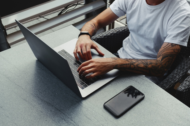Jóképű fickó tetovált kézzel öltözött fehér inget dolgozik laptop, férfi gépelés szöveget notebook billentyűzet. Szabadúszó dolgozik az új indítási projekt a városi kávézóban, ingyenes vezeték nélküli övezet - Fotó, kép