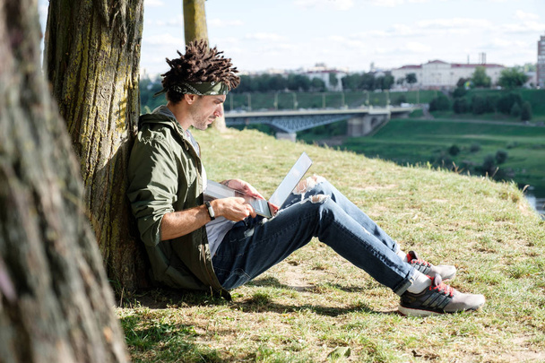 Young millennial student with dreadlocks sitting on grass and using portable laptop computer outdoor in public park. Freelancer using notebook for distance job, remote working, e-learning - Foto, Imagem