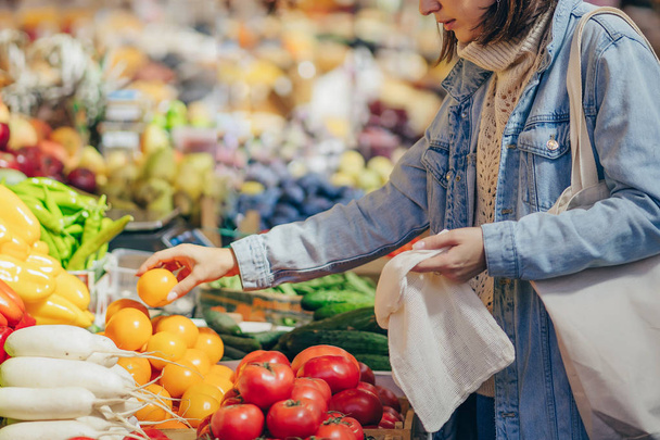 女性は食品市場で果物や野菜を選択します。買い物に再利用可能なエコバッグ。持続可能なライフスタイル。環境にやさしいコンセプト.  - 写真・画像