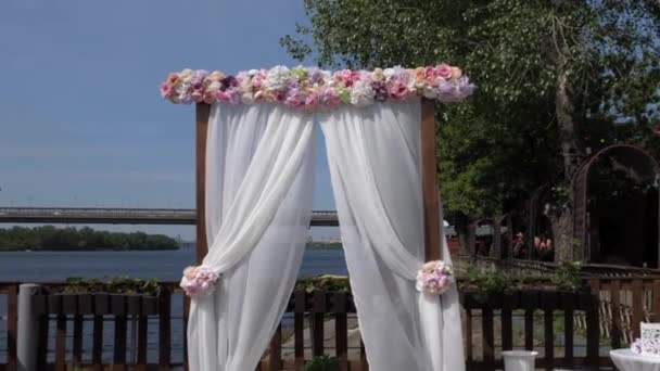 Весільна арка з білої тканини і дерев'яна основа на берегах Дніпра на фоні мосту. Весільна церемонія, стільці влаштовуються для гостей. - Кадри, відео