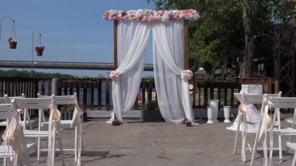橋の背景にドニエパー川のほとりに白い生地と木製のベースの結婚式のアーチ。結婚式、椅子を手配. - 映像、動画
