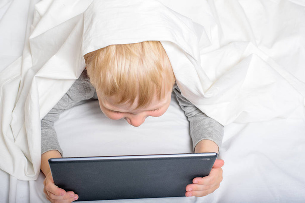 Little blond boy is looking at something on a tablet. Lying in bed and hiding under the covers. Gadget Leisure. Top view - Photo, Image