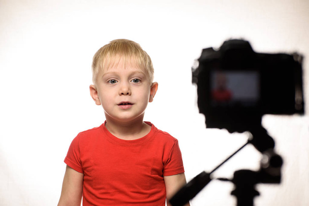 Rapaz loiro sério está a gravar um vídeo na câmara DSLR. Um pequeno blogueiro de vídeo. Fundo branco
 - Foto, Imagem