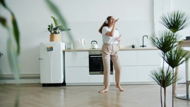 Slow motion of cute girl in headphones singing in spoon in kitchen dancing alone - Záběry, video