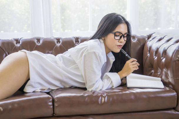 Sexy zakenvrouw of secretaris zet glazen lezing dagboek dragen van wit overhemd grote borst zittend rusten op Bank werk thuis - Foto, afbeelding