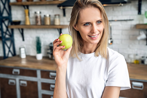 Close up of a beautiful blond who smiles and holds an apple in her hand. girl stands in the kitchen in a white t shirt - Foto, Imagem