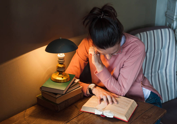 Diligent student. A tired girl student leaned over a thick book with many bookmarks, at a tiny table, under a warm lamp. - Φωτογραφία, εικόνα