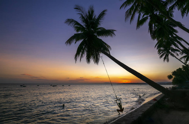 Silueta dětí se houpá na kokosovém stromě a při západu slunce na nádherné pláži - Fotografie, Obrázek