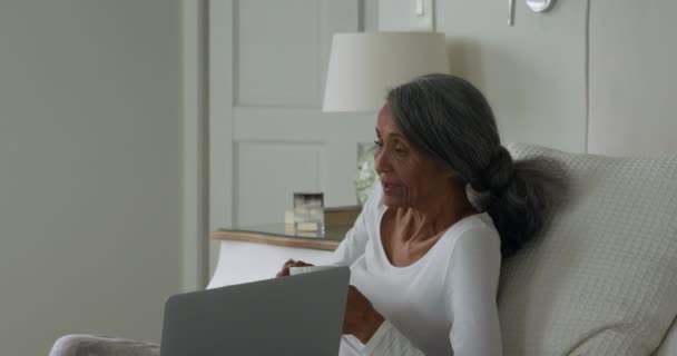 Side view of a mature mixed race couple with a laptop computer sitting on a bed at home talking - Imágenes, Vídeo