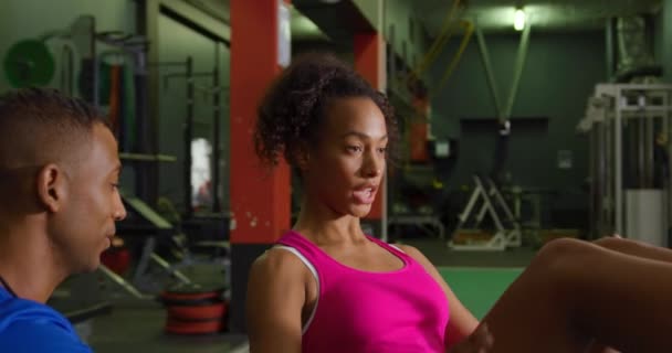Side view close up of a young mixed race woman exercising in a gym with a young mixed race male personal trainer, preparing to do crunches - Footage, Video