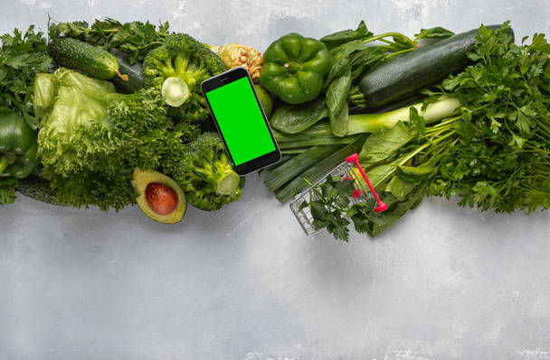 Smartphone with blank screen and green vegetables on light background with shopping trolley. Food ordering through mobile cell phone application concept - Foto, Imagen