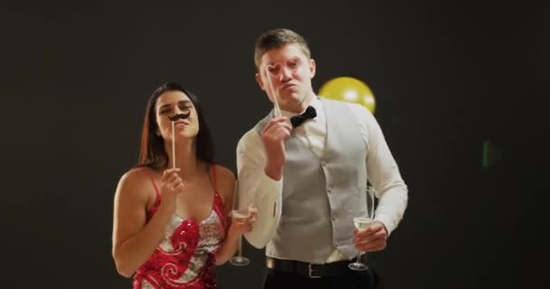 Μπροστινή όψη κοντά σε ένα έξυπνα ντυμένο χαμόγελο νεαρό καυκάσιο ζευγάρι πίνοντας ποτήρια σαμπάνιας, χορεύοντας κάτω από τα μπαλόνια που πέφτουν και ποζάρισμα για μια εικόνα με σκηνικά φωτογραφίες κόμμα σε ένα κόμμα, σε μαύρο φόντο - Πλάνα, βίντεο
