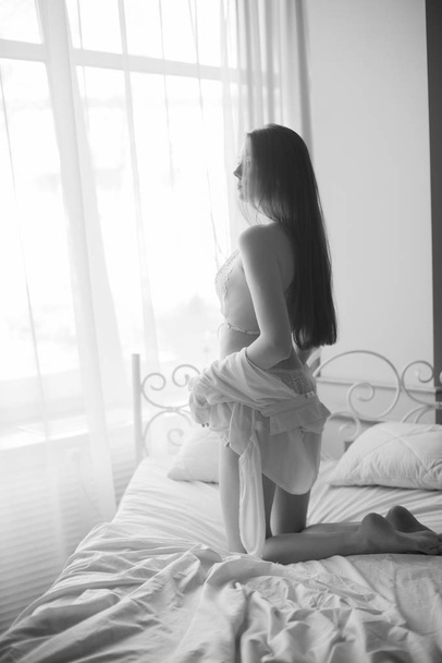 Young attractive girl with long dark hair in lace white underwear and a white silk shirt by the bed. Boudoir photo of a young girl - Φωτογραφία, εικόνα