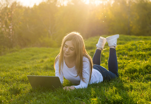 Young beautiful woman with a laptop lies on the grass in sunset. - Photo, Image