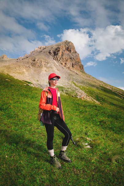 Girl athlete photographer in a red cap sunglasses and a yellow backpack stands on a green slope against the background of the epic cliffs of the Caucasus - Photo, image