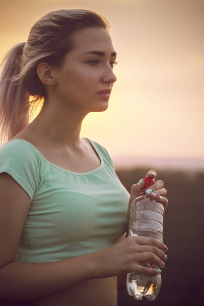Silhouettenprofil eines schönen Mädchens mit einer Flasche Wasser nach dem Joggen auf einem Feld bei Sonnenuntergang, Frau freut sich auf die Natur, Konzeptsport und Gesundheit - Foto, Bild