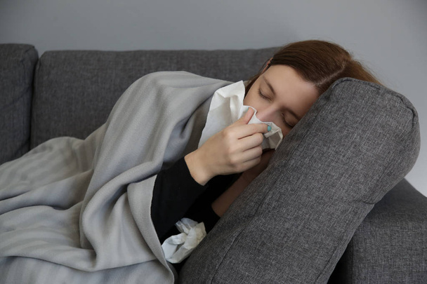 Młoda kobieta na kanapie dmuchanie nosa w białą tkankę papierową. Studio strzał młodej kobiety z objawami alergii kichanie do tkanek. Grypa, zimno lub objawy alergii.  - Zdjęcie, obraz