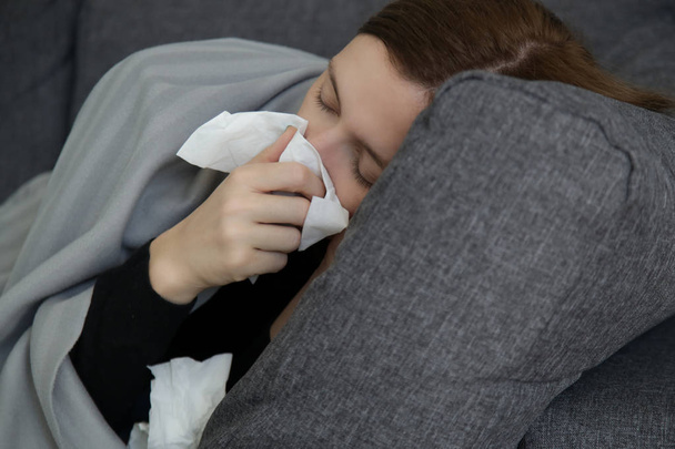 Young woman on the sofa blowing nose into a white paper tissue. Studio shot of young woman with allergy symptoms sneezing into a tissue. Flu, cold or allergy symptom.  - Foto, Bild