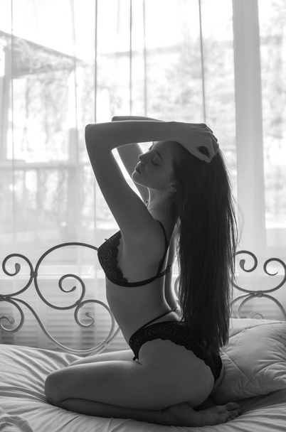 Young attractive woman with long dark hair in black lace lingerie on a bed. Boudoir photo of a young girl - Фото, изображение