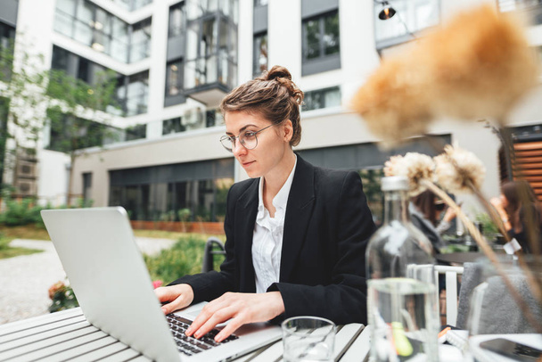 Young business woman sitting at coffee shop on veranda and working on laptop. Portrait of female freelancer wearing glasses with computer - Фото, зображення