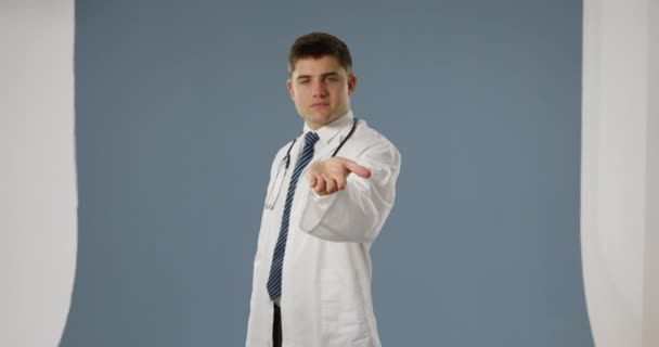 Front view of young Caucasian male doctor smiling to camera with his hand held out and palm up on a grey background - Video