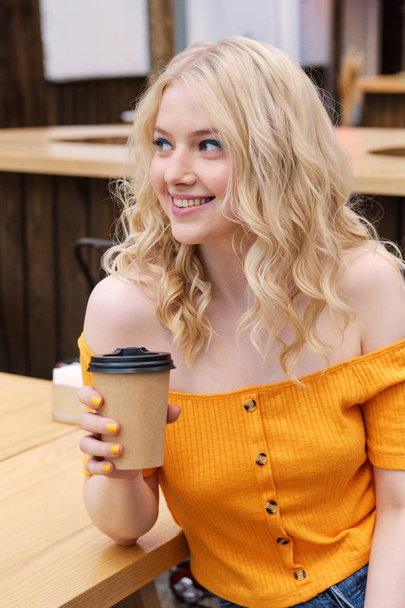 Attractive smiling casual blond girl happily looking away with coffee to go in hand in courtyard of cafe   - Foto, imagen