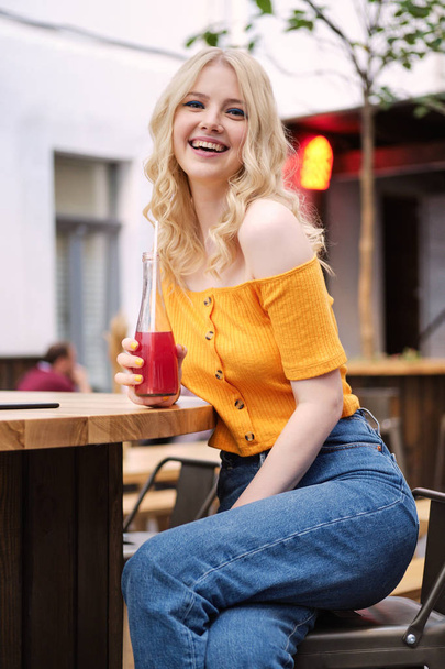 Dość radosna blond dziewczyna szczęśliwie patrząc w aparacie z lemoniadą odpoczynku na dziedzińcu kawiarni - Zdjęcie, obraz