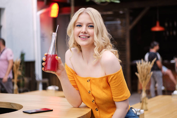 Piękna uśmiechnięta blond dziewczyna szczęśliwie patrząc w aparacie z lemoniadą spoczywającą na dziedzińcu kawiarni - Zdjęcie, obraz