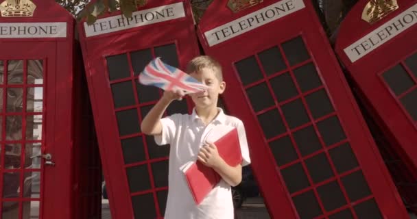 Αργή κίνηση πορτρέτο του χαριτωμένο αγόρι κουνώντας βρετανική σημαία στέκεται έξω μόνο χαμογελώντας κοιτάζοντας την κάμερα. Στο φόντο των αγγλικών κόκκινων τηλεφωνικών θαλάμων. Ταξίδι έννοια. - Πλάνα, βίντεο