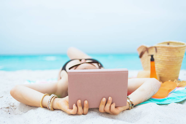 Seen from behind elegant middle age woman with long curly hair in elegant black swimsuit with book while laying on a white beach. - Valokuva, kuva