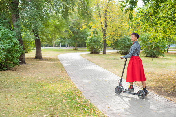 A young woman with purple hair rides an electric scooter in a park. A stylish girl with a shaved temple in a plaid shirt, a long red skirt and a bow tie is riding around the city on a modern device.  - Foto, imagen