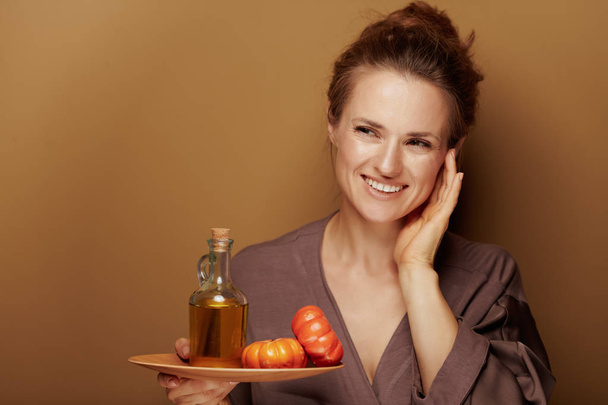 Hello autumn. Portrait of smiling young woman in a bathrobe showing pumpkin oil and touching face and looking at copy space against beige background. - Photo, Image