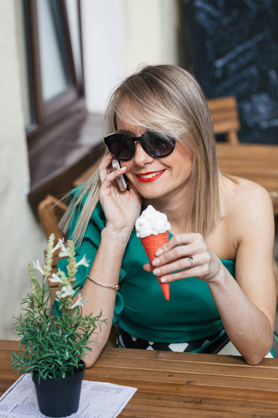 portrait of attractive blonde young woman in sunglasses, green dress and with red lipstick sits at table of street cafe. Woman holding red ice cream cone and calling by phone - Fotoğraf, Görsel