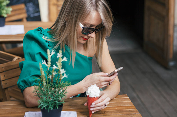portrait of attractive blonde young woman in sunglasses, green dress and with red lipstick sits at table of street cafe. Woman holding red ice cream cone and looking at cell phone - Foto, immagini