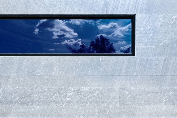 Moderne gevel van grijze stenen platen met een langwerpig raam dat de lucht weerspiegelt - Foto, afbeelding
