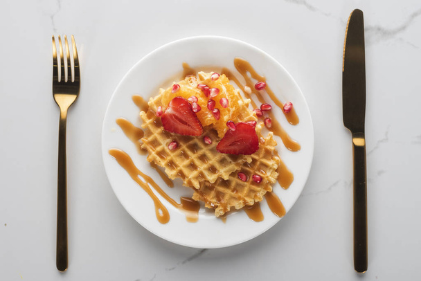 waffles com morangos em uma placa branca em um fundo branco com garfo dourado e faca
 - Foto, Imagem