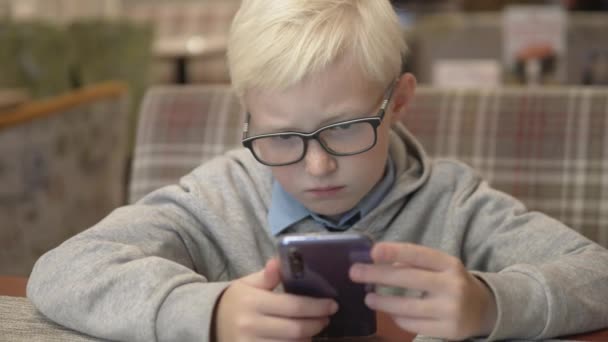 Egy komoly fiú szemüveges ül egy kávézóban, és játszik a mobiltelefonján - Felvétel, videó