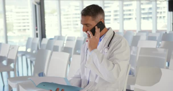 Close up side view of a middle aged Caucasian male doctor sitting in a chair at an empty seminar holding a file and talking on a smartphone - Video