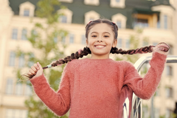 Having hair in two plaits. Adorable girl holding pigtails hair on urban background. Small cute girl with long brunette hair outdoor. Little hair model with beauty look - Valokuva, kuva