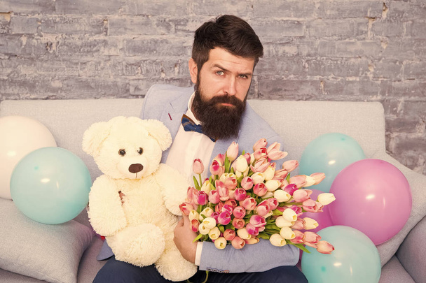 How successfully ask her dating. Romantic man with flowers and teddy bear sit on couch waiting girlfriend. Romantic gift. Macho ready romantic date. Man wear blue tuxedo bow tie hold flowers bouquet - Foto, afbeelding