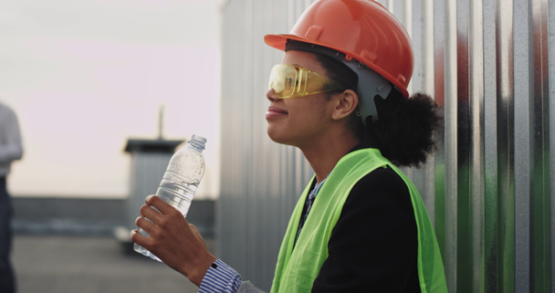 Thirsty lady African ethnic drinking water at construction site from a plastic bottle she wearing safety equipment helmet and yellow glasses - Imágenes, Vídeo