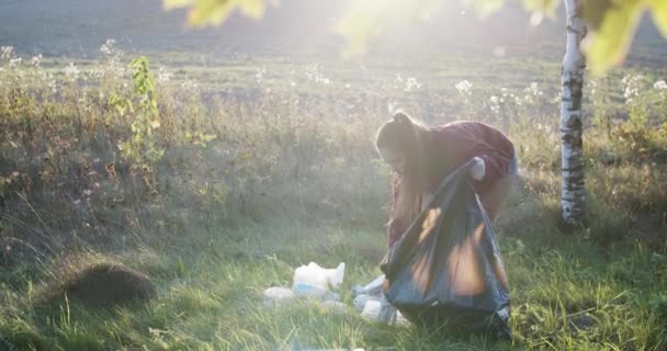 junge Frau sammelt Müll im Freien Natur Hintergrund Zeitlupe. Mädchen sammeln freiwillig recycelbaren Plastikmüll in der sonnigen Landschaft. Ökologischer Aktivismus freiwillige Erdpflege - Filmmaterial, Video
