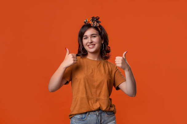 Hezká brunetka v oranžovém tričku a hlavni, která drží palce, vyjadřuje pozitivní hodnocení v izolaci nad oranžovým pozadím. Líbí se mi to! - Fotografie, Obrázek