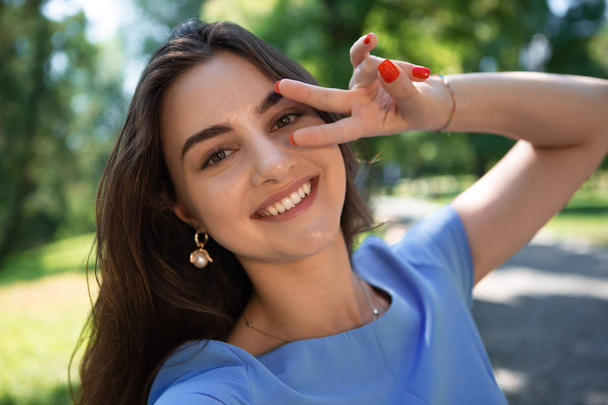 Mavi elbiseli gülümseyen genç kadın parkta oturuyor ve akıllı telefonla selfie çekiyor. - Fotoğraf, Görsel