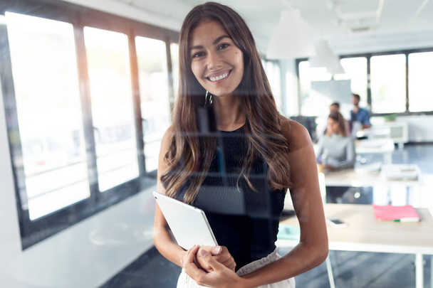 Smiling young businesswoman looking at camera while standing and holding her digital tablet in the modern startup office. - Foto, afbeelding