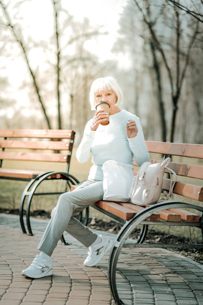 Пожилая мадам с кофе. Пожилая дама в повседневной одежде и белых кроссовках пьет кофе, чтобы попасть на скамейку запасных.
. - Фото, изображение