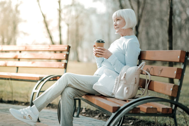 Blue-rinsed woman. Charming blue-rinsed woman with short haircut concernedly sitting with paper cup on the bench - Fotografie, Obrázek