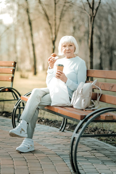 Fascinating beaming senior female eating cookies and drinking coffee - Zdjęcie, obraz