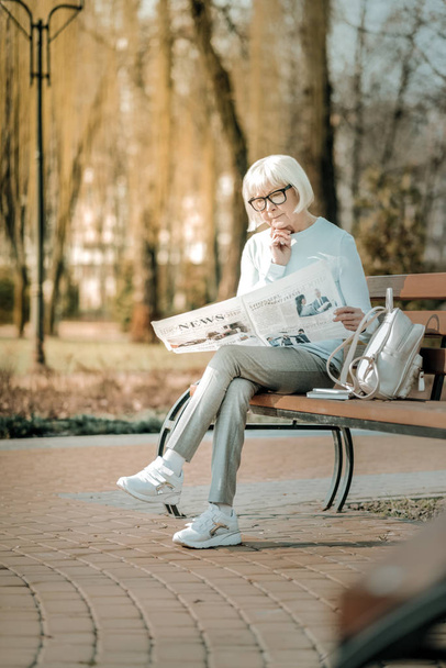 Pleasant superannuated lady in glasses thoughtfully reading newspaper on bench - Photo, image