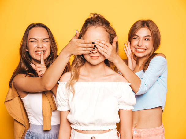 Três jovens bonitas meninas hipster sorridentes em roupas de verão na moda.Sexy mulheres despreocupadas posando perto da parede amarela no studio.Modelos surpreendendo seus amigos.Eles cobrem os olhos e abraçando por trás
 - Foto, Imagem